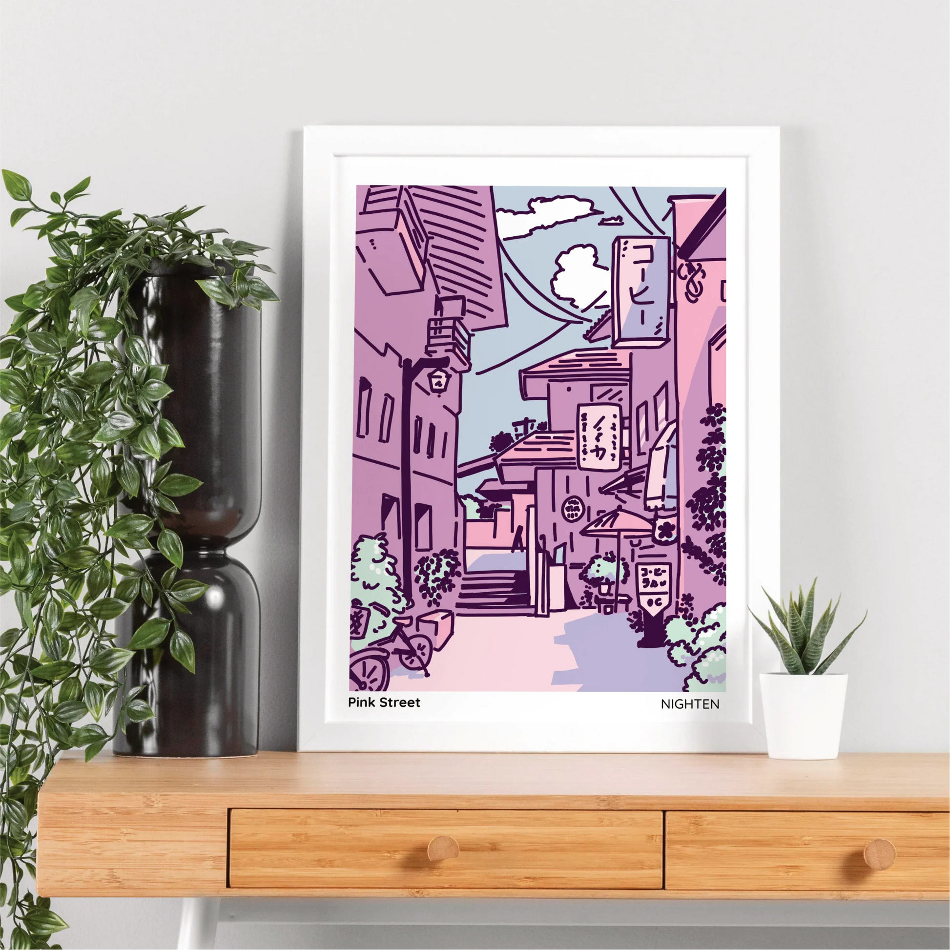Framed artwork of a stylized cityscape on a wooden surface with plants.